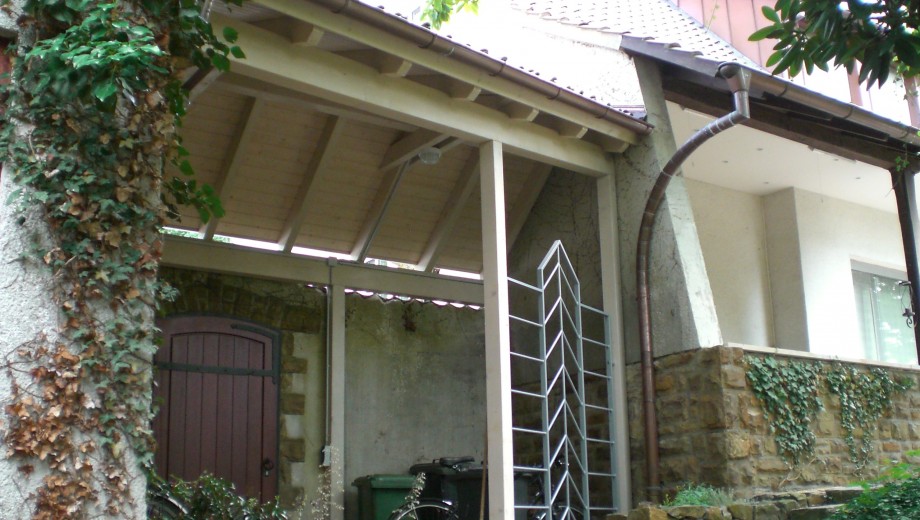Überdachter Bereich mit Holzregal, dunkler Tür und Efeu an den Wänden; ruhige, natürliche Atmosphäre.