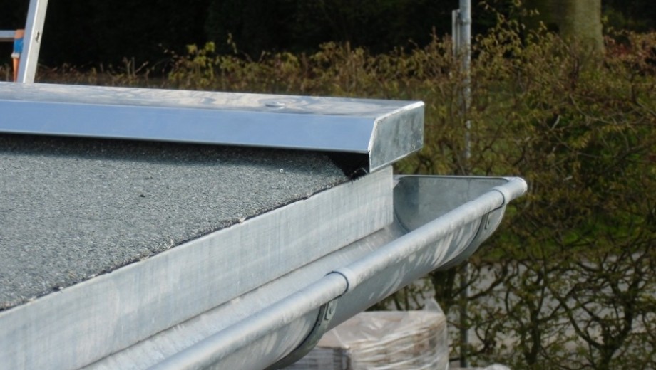 Ein graues Kiesdach mit silberner Metallregenrinne vor unscharfem, grün-braunem Hintergrund.