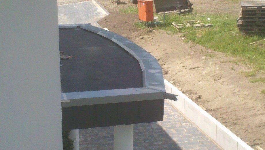 Baustelle mit Flachdach, orangefarbenem Container, Erdbereich und Baumaterialien im Hintergrund.