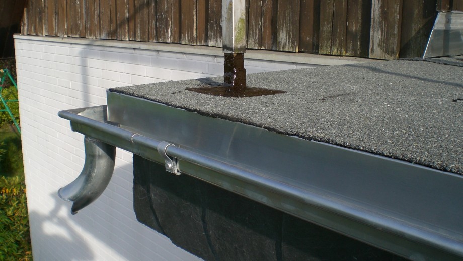 Ein Flachdach mit Kies, Regenwasser fließt aus einem Rohr in eine metallene Regenrinne.