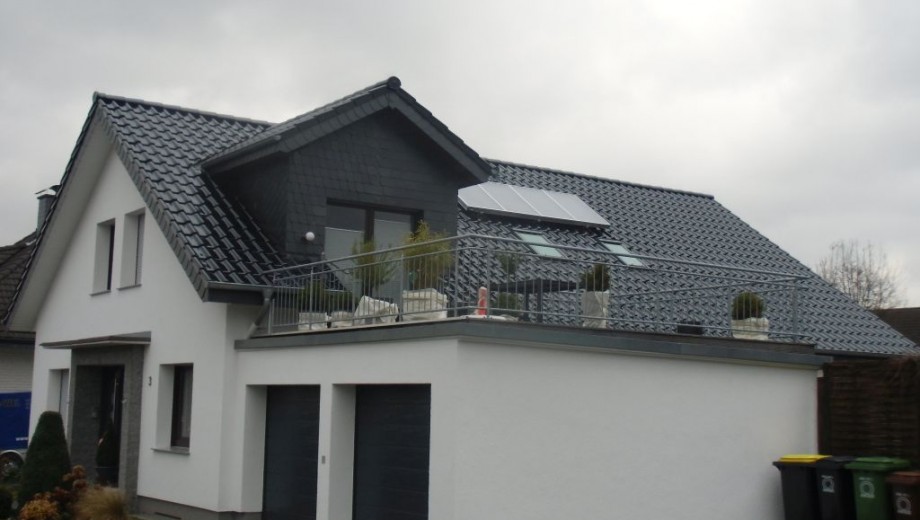 Modernes Einfamilienhaus mit Satteldach, Solarpanelen und Dachterrasse, umgeben von Pflanzen.