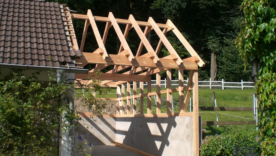 Baustelle mit Holzrahmen für Dach, umgeben von Bäumen, neben Gebäude mit Ziegeldach. Ruhige Atmosphäre.