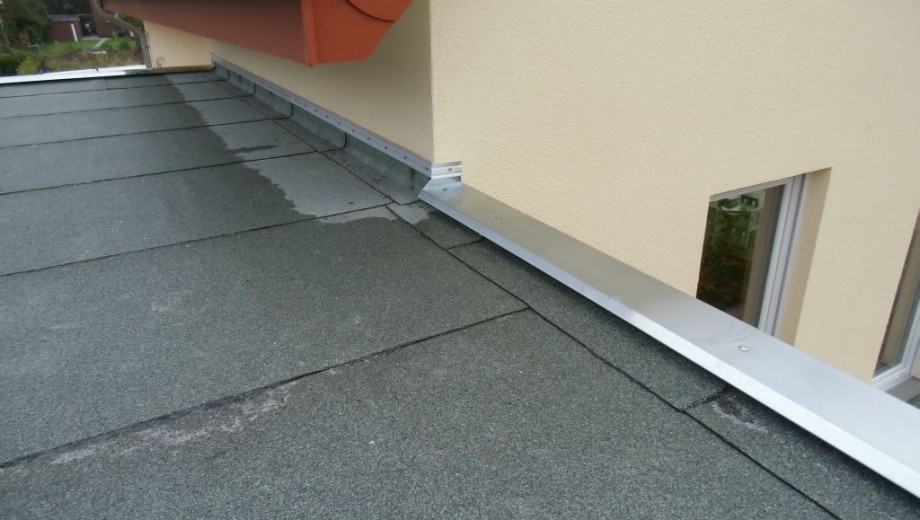 Ein Flachdach mit dunklen Dachbahnen, silberner Metallleiste und beiger Wand mit Fenster.