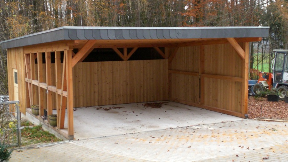 Holz-Carport mit Betonboden, herbstliche Bäume im Hintergrund, kleines Fahrzeug daneben.