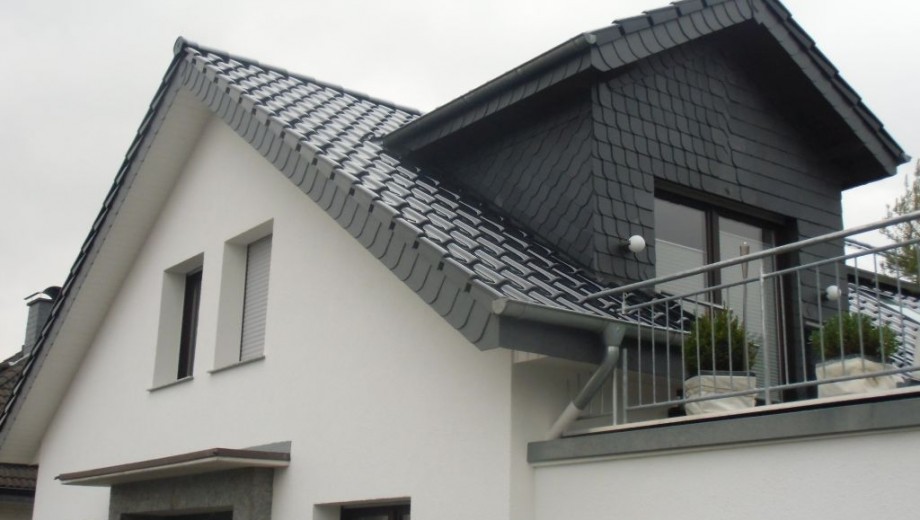 Modernes Haus mit geneigtem Dach, Dachfenstern und Balkon; weiße Fassade, graues Dach.