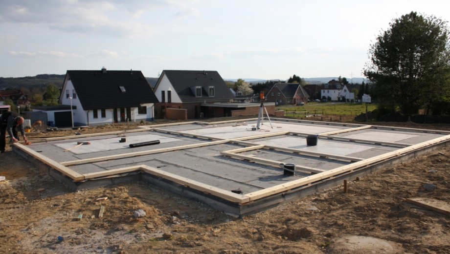 Baustelle mit Betonfundament, Holzbalken und zwei Arbeitern in einem Wohngebiet. Fertige Häuser im Hintergrund.