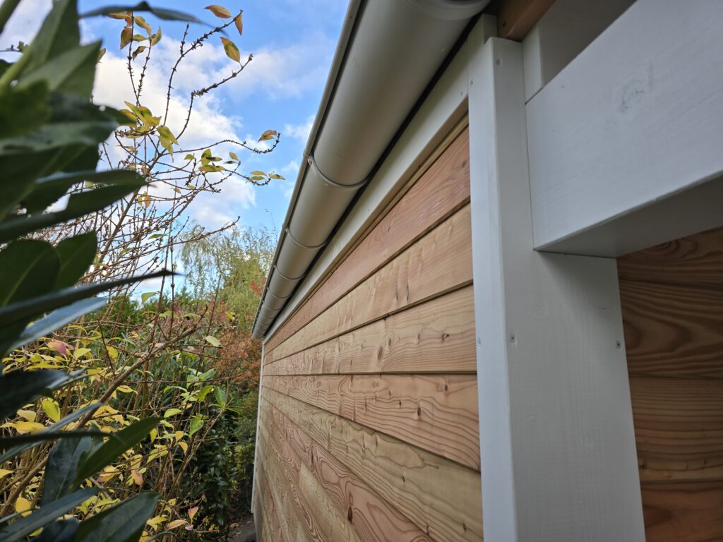 Holzwand mit Dachrinne, umgeben von Herbstpflanzen unter blauem Himmel mit weißen Wolken.