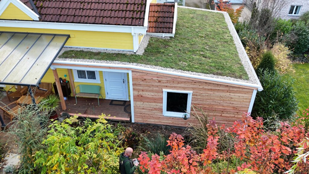 Ein Haus mit Holz- und Gelbfassade, begrüntem Dach, Herbstpflanzen und einem Mann im Garten.