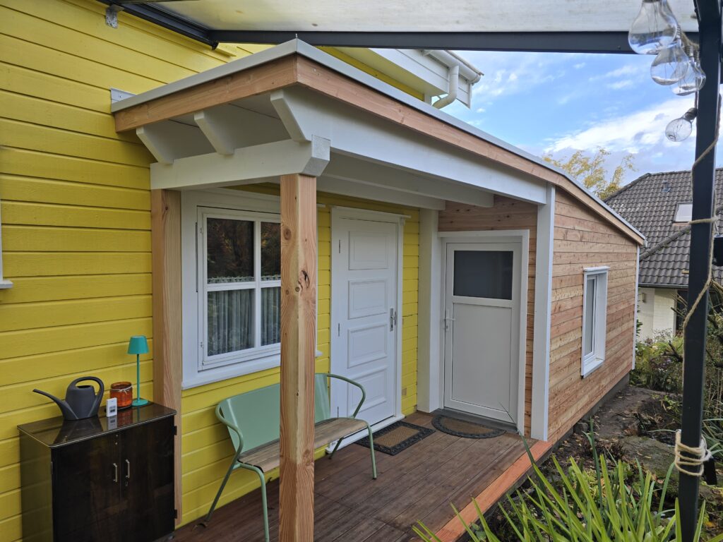 Ein gelbes Haus mit weißer Tür, grünem Stuhl und kleinem Tisch im einladenden Eingangsbereich.