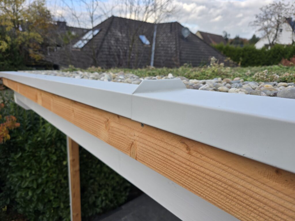 Ein begrüntes Flachdach mit Pflanzen und Kieselsteinen, umrahmt von einer Metallkante, unter blauem Himmel.