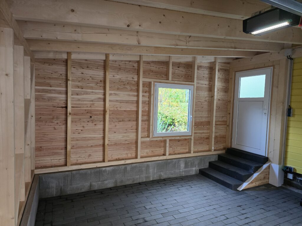 Innenraum einer Garage mit Holzpaneelwänden, grauem Pflasterboden, kleinem Fenster und weißer Tür.