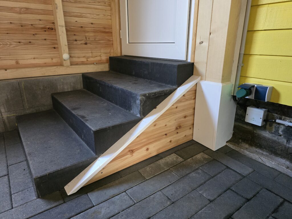 Dunkle Steintreppe führt zu weißer Tür, umgeben von Holzverkleidung und gelber Wand.