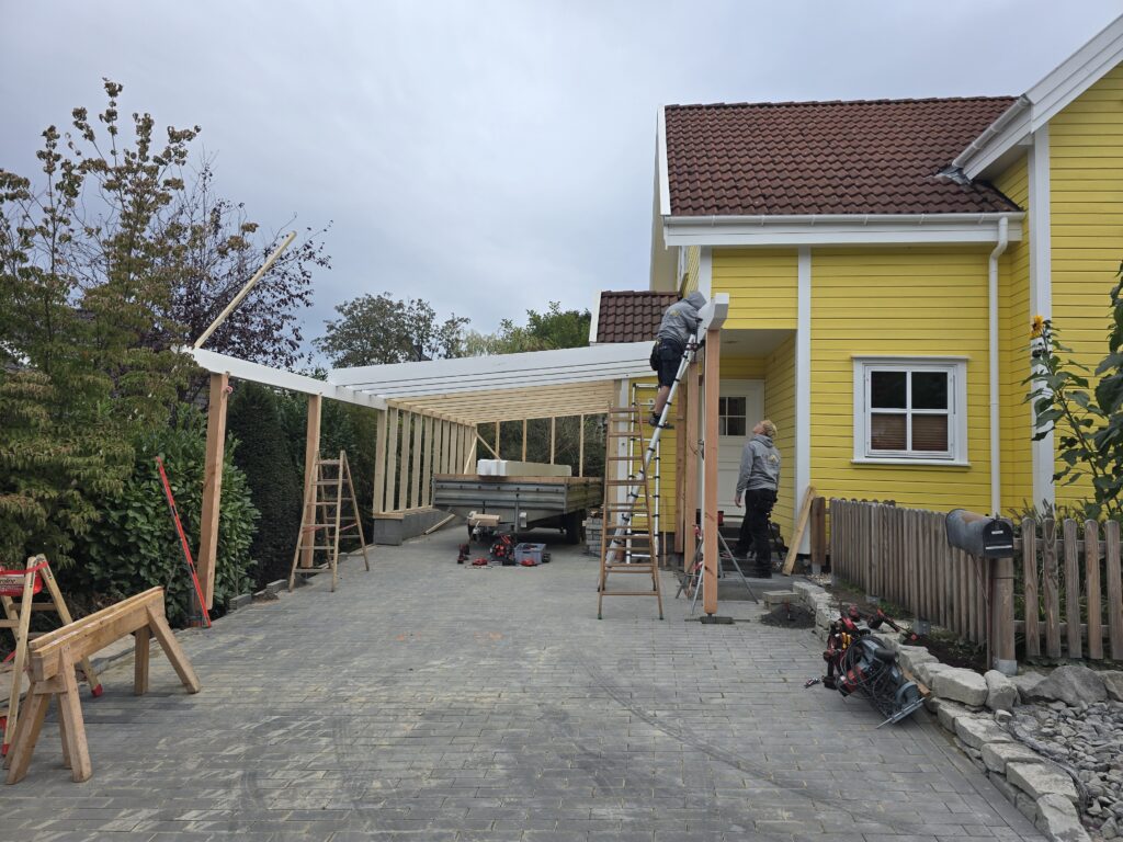 Zwei Personen bauen einen hölzernen Carport vor einem gelben Haus mit rotem Ziegeldach.