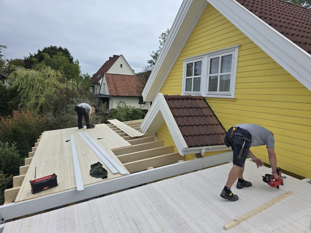 Zwei Personen arbeiten konzentriert auf einer Holzplattform an einem gelben Haus mit rotem Ziegeldach.