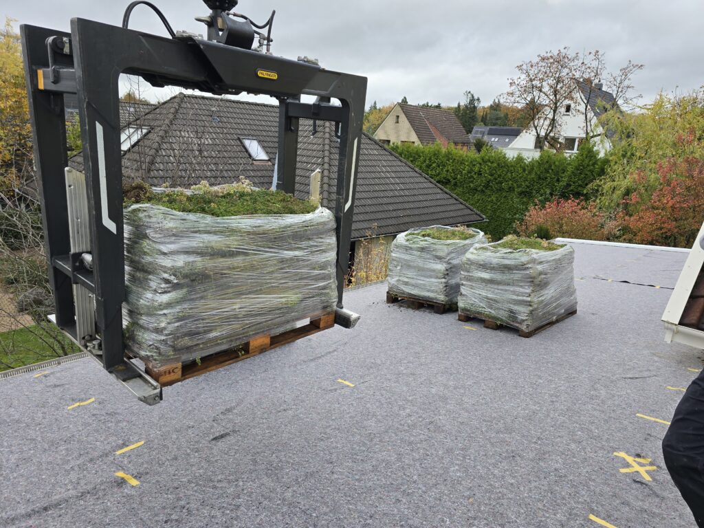 Ein Kran hebt Paletten mit grüner Vegetation auf ein graues Dach in herbstlicher Umgebung.