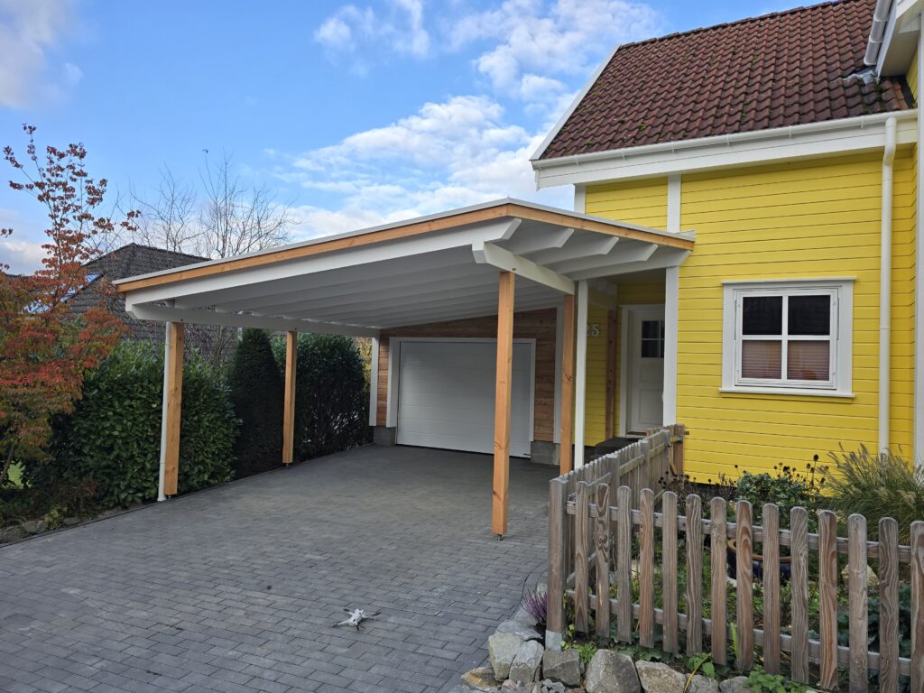 Ein gelbes Haus mit rotem Ziegeldach, Carport, Holzzaun und herbstlich gefärbtem Baum davor.