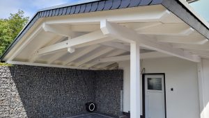 Moderner Carport mit weißem Dach, Holzbalken, Gabionenwand und weißer Tür, sonniger Tag.