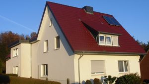 Ein zweistöckiges Haus mit rotem Dach und Solarpanel, umgeben von Bäumen, bei sonnigem Wetter.